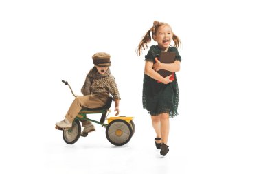 Portrait of cheerful girl with books playing with little boy on small bike isolated over white background. Concept of fashion, childhood, game, school, fun, education. Copy space for ad