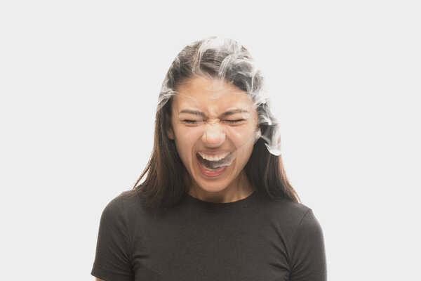 Shouting young girl with dark hair releases smoke from the mouth isolated over white background. Concept of mental health, art, human emotions, challenges, ad