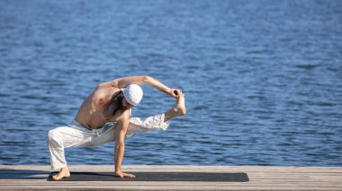Peace and quiet. Summer morning. Young man practicing yoga on river pier at sunrise, outdoors. Concept of mental health, harmony with nature, emotions, skills. Wellness and wellbeing