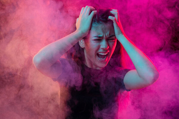 Emotional pain. Young dark hair woman shouting isolated over neoned pink background with clouds of smoke. Concept of mental health, art, human emotions, challenges, ad
