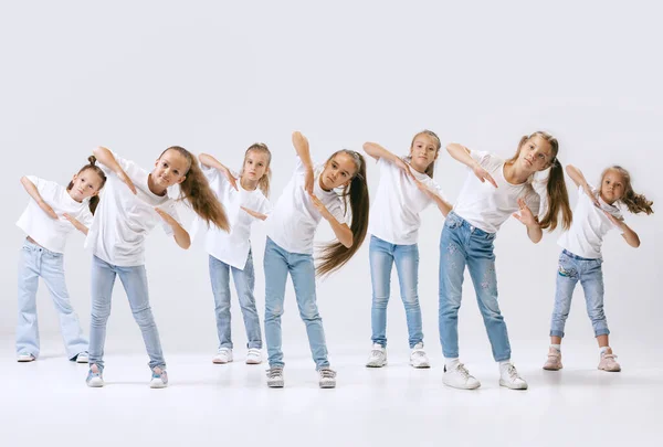 Modern choreography lesson. Dance group of happy, active little girls in jeans and t-shirts dancing isolated on white studio background. Concept of music, fashion, art, childhood, hobby.