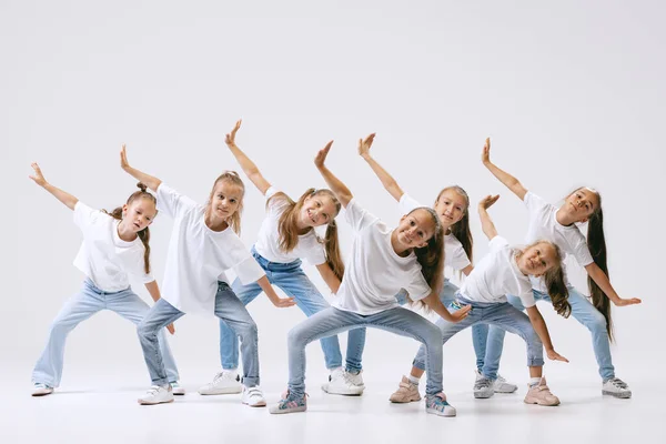 Modern choreography lesson. Dance group of happy, active little girls in jeans and t-shirts dancing isolated on white studio background. Concept of music, fashion, art, childhood, hobby.