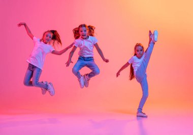 Group of school age girls, cheerful kids in casual style clothes dancing, jumping isolated on pink background in yellow neon light. Hip-hop dance, street style. Concept of music, fashion, art