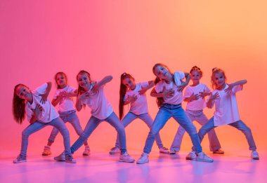 Group of school age girls, cheerful kids in casual style clothes dancing, jumping isolated on pink background in yellow neon light. Hip-hop dance, street style. Concept of music, fashion, art