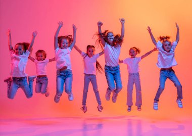 Modern contemp dance. Happy kids, school age girls in white t-shirts and jeans jumping isolated on pink background in yellow neon light. Concept of music, fashion, art, childhood
