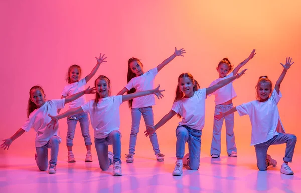 Hip-hop dance, street style. Group of children, school age girls in casual style clothes dancing in choreography class isolated on pink background in yellow neon light. Concept of music, fashion, art