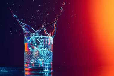 Bursts of water. Whiskey glass with ice cubes and water splashes standing on mirror surface over gradient black orange background in neon light. Concept of art, beauty, drinks. Copy space for ad