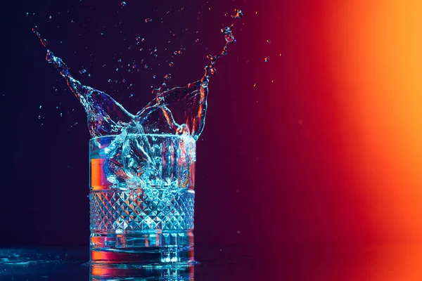 Bursts of water. Whiskey glass with ice cubes and water splashes standing on mirror surface over gradient black orange background in neon light. Concept of art, beauty, drinks. Copy space for ad