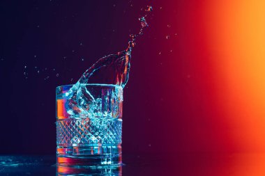 Vision. Whiskey glass with ice cubes and water splashes standing on mirror surface over gradient black orange background in neon light. Concept of art, beauty, drinks. Copy space for ad