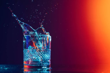 Closeup glass with ice cubes and water splashes standing on mirror surface over gradient black orange background in neon light. Concept of art, beauty, drinks. Copy space for ad