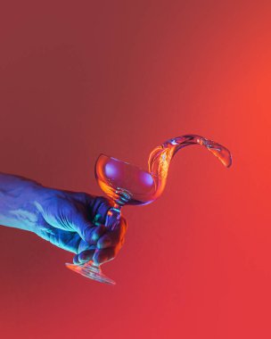 Mesmerizing effect. Male hand pouring clean fluid from cocktail glass over bright dark red background in neon light. Concept of art, beauty, fashion, drinks, creativity. Copy space for ad