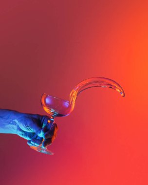 Flying water. Male hand pouring clean fluid from cocktail glass over bright dark red background in neon light. Concept of art, beauty, fashion, drinks, creativity. Copy space for ad