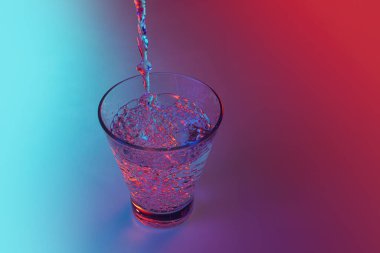 Composition with water flows into glass placed on table over gradient multicolored background in neon light. Bar, drinks, health and art. Copy space foe ad, text.