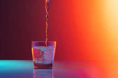 Composition with water flows into glass placed on table over gradient multicolored background in neon light. Bar, drinks, health and art. Copy space foe ad, text.