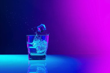 Ice cube falls into a transparent glass of water standing on mirror surface over gradient blue-pink background in neon light. Concept of art, beauty, drinks. Copy space for ad