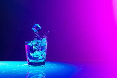 Ice cube falls into a transparent glass of water standing on mirror surface over gradient blue-pink background in neon light. Concept of art, beauty, drinks. Copy space for ad