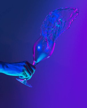 Greeting toast. Authentic hand pouring pure liquid from wine or champagne glass over bright blue gradient background in neon light. Art, drinks, creativity