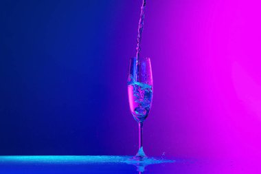 Flying fluid. Authentic hand pouring pure water splash from wine or champagne glass over bright blue-pink gradient background in neon light. Art, drinks, creativity