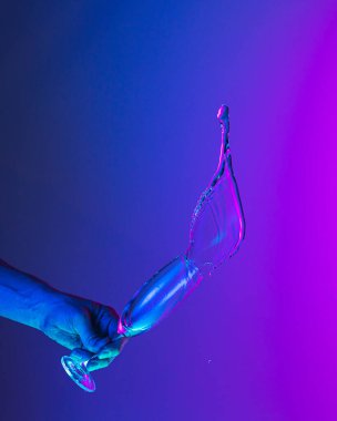 Flying fluid. Authentic hand pouring pure water splash from wine or champagne glass over bright blue-pink gradient background in neon light. Art, drinks, creativity