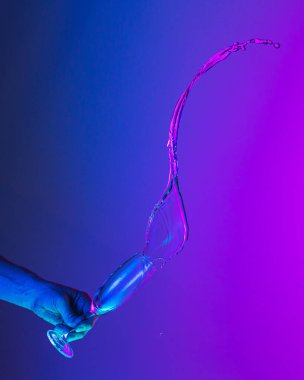 Greeting toast. Authentic hand pouring pure liquid from wine or champagne glass over bright blue-pink gradient background in neon light. Art, drinks, creativity
