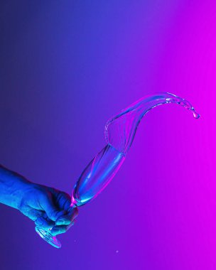 Flying fluid. Authentic hand pouring pure water splash from wine or champagne glass over bright blue-pink gradient background in neon light. Art, drinks, creativity