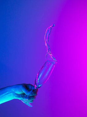 Greeting toast. Authentic hand pouring pure liquid from wine or champagne glass over bright blue-pink gradient background in neon light. Art, drinks, creativity