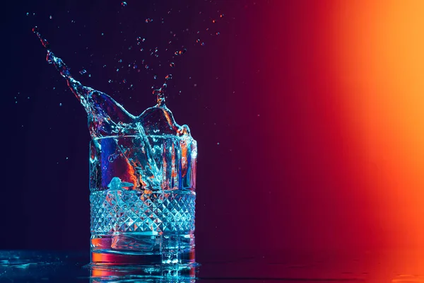 Closeup glass with ice cubes and water splashes standing on mirror surface over gradient black orange background in neon light. Concept of art, beauty, drinks. Copy space for ad