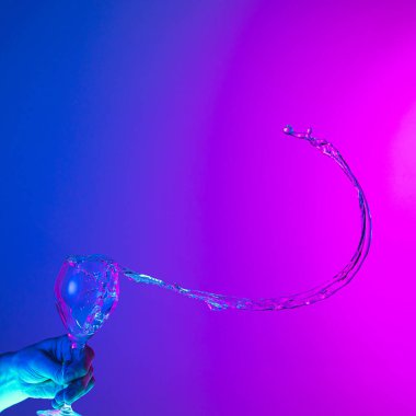 Greeting toast. Authentic hand pouring pure liquid from wine or champagne glass over bright blue-pink gradient background in neon light. Art, drinks, creativity