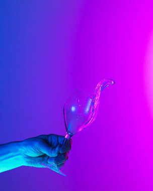 Greeting toast. Authentic hand pouring pure liquid from wine or champagne glass over bright blue-pink gradient background in neon light. Art, drinks, creativity