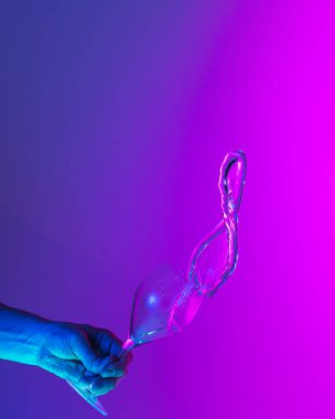 Flying fluid. Authentic hand pouring pure water splash from wine or champagne glass over bright blue-pink gradient background in neon light. Art, drinks, creativity