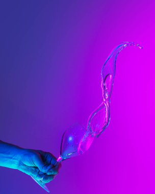 Greeting toast. Authentic hand pouring pure liquid from wine or champagne glass over bright blue-pink gradient background in neon light. Art, drinks, creativity
