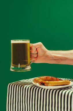 Beer and fried frankfurter. Human hand with mug of cold frothy beer over dark green background. Oktoberfest, vacation, happiness, festival and ad concept. Retro style.