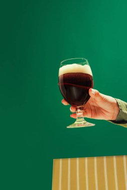 Tasting beer. Female hand holding glass of cold dark beer over dark green background. Oktoberfest, vacation, happiness, drinks, Bavaria, travel, fest and ad concept. Retro style.