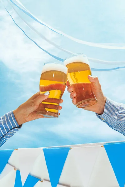 Closeup male and female hands with glass of beer toasting isolated over blue white background with Oktoberfest decoration. Sport, fan, bar, pub, celebration, soccer football concept.