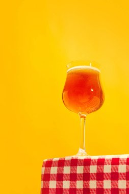 Glass of cold frothy light beer standing on table with plaid cloth over bright yellow background. Oktoberfest, vacation, happiness, drinks, travel, fest and ad concept. Retro style.