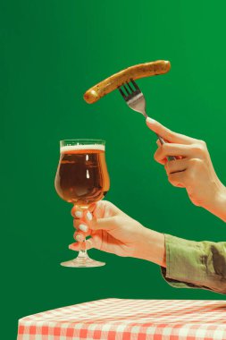 Bon appetit. Female hand with glass of cold light beer and fried sausage over green background. Oktoberfest, vacation, happiness, drinks, beach, travel, fest and ad concept. Retro style.