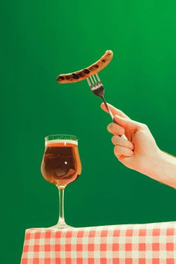 Bon appetit. Female hand with glass of cold light beer and fried sausage over green background. Oktoberfest, vacation, happiness, drinks, beach, travel, fest and ad concept. Retro style.