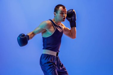 Practicing punch. Muscled man, male boxer in blue uniform and boxing gloves training isolated on navy background in neon. Strength, attack and motion concept. Copy space for ad