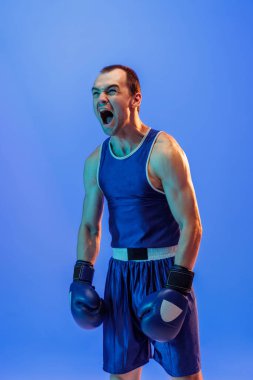 Winner emotions. Male boxer in blue uniform and boxing gloves shouting isolated on blue background in neon. Strength, sport, facial expression and motion concept. Copy space for ad