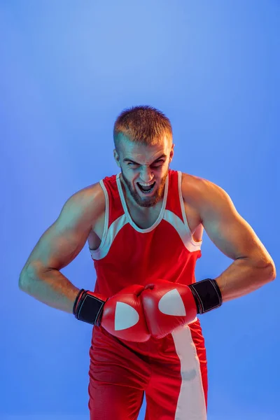 Winner emotions. Sporty man during boxing exercises. Male boxer in red uniform and boxing gloves training isolated on blue background in neon. Strength, attack and motion concept. Copy space for ad