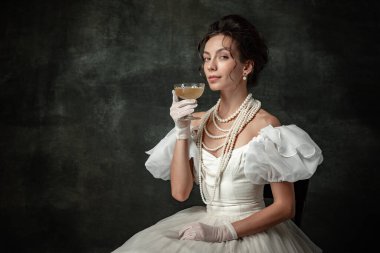 Tasting red wine. Portrait of young charming girl in image of medieval royal person or viscountess posing isolated on dark vintage background. Comparison of eras concept, fashion, art, ad.