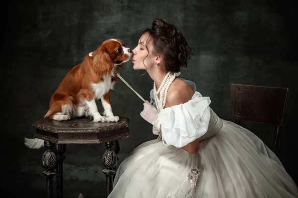 Kiss. Vintage portrait of young elegant woman in image of medieval person in renaissance style dress with little cute dog isolated on dark background. Comparison of eras, beauty, history, art