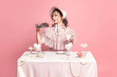 Tea ceremony. Young pretty woman with retro style hairdo in bonnet and vintage dress sitting at table isolated on pink background. Eras comparison, art, beauty holiday, fashion, emotions, and ad
