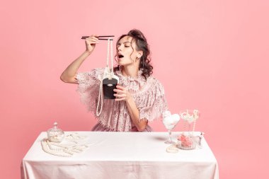 Creative image with young beautiful girl with retro style hairdo wearing medieval dress sitting at served table isolated on pink background. Eras comparison, art, beauty, fashion, emotions, and ad