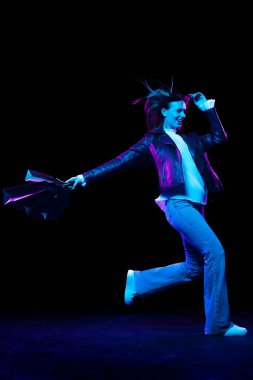 In a hurry to shop. Fashionable stylish happy woman with shopping bags running isolated over dark background in neon light. Shopping, sales, ad, black friday, New Year and Christmas concept