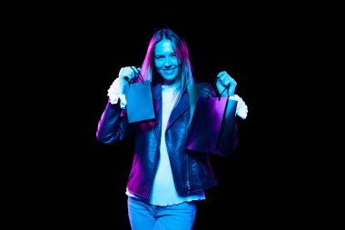 Long-awaited purchase. Portrait of young excited woman with shopping bags isolated over dark background in neon light. Shopping, sales, ad, black friday, seasonal sales concept