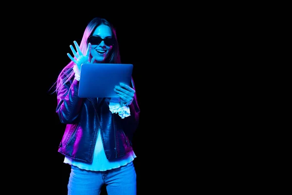 Greets friend in online meeting. Fashionable young pretty girl using digital tablet isolated on dark background in neon light. Concept of emotions, fashion, style, sales, youth, aspiration.
