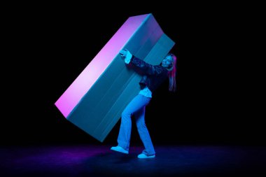 Long-awaited purchase. Young excited woman carries huge carton box isolated over dark background in neon light. Shopping, sales, ad, black friday, New Year and Christmas concept