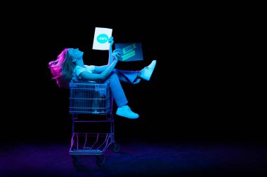 Cheerful astonished girl sitting in shopping cart and inviting for shopping, black friday, seasonal sales isolated over dark background in neon light. Cyber monday and online purchases.