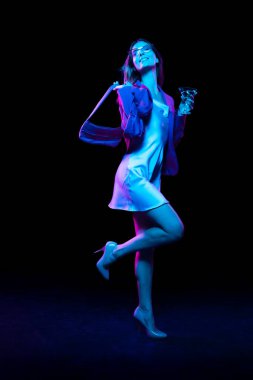 Party time. Young fashionable stylish happy woman tasting cocktail isolated over dark background in neon light. Shopping, sales, ad, black friday, New Year and Christmas concept